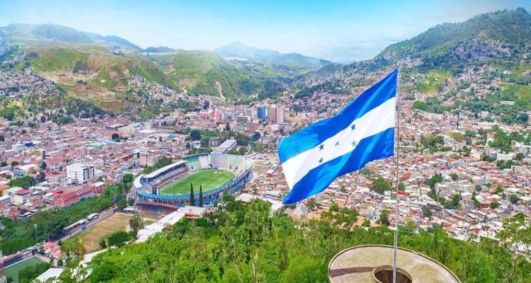 Large Honduran flag waves above a sprawling valley city with stadiums and mountains in the distance.