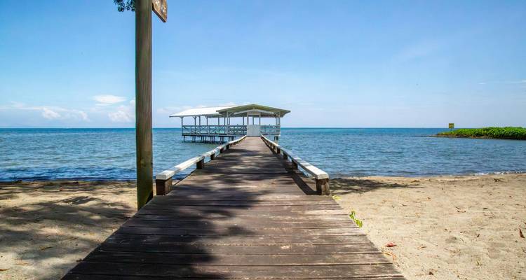 Wooden boardwalk stretches out to a small pavilion over clear blue water beside a sandy shore.