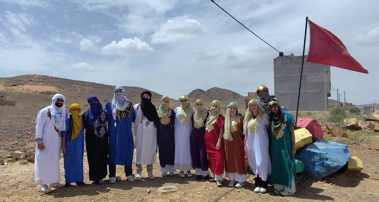 Grand groupe de touristes vêtus de robes traditionnelles colorées posant dans un décor de désert rocheux à côté d'un drapeau marocain.