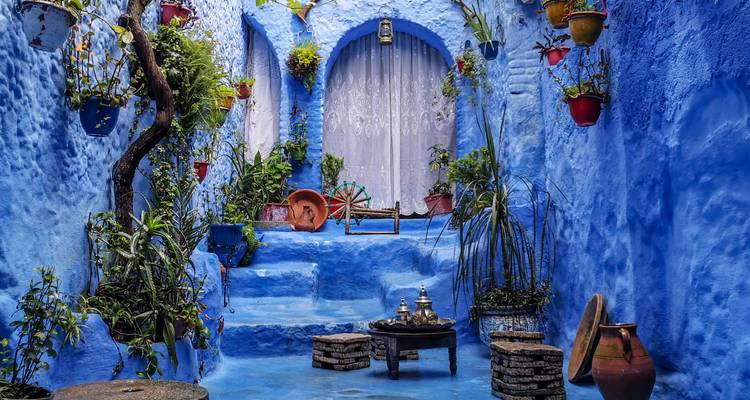 Charmante cour bleue avec des plantes en pot, une arche et un service à thé marocain traditionnel sur des tabourets.