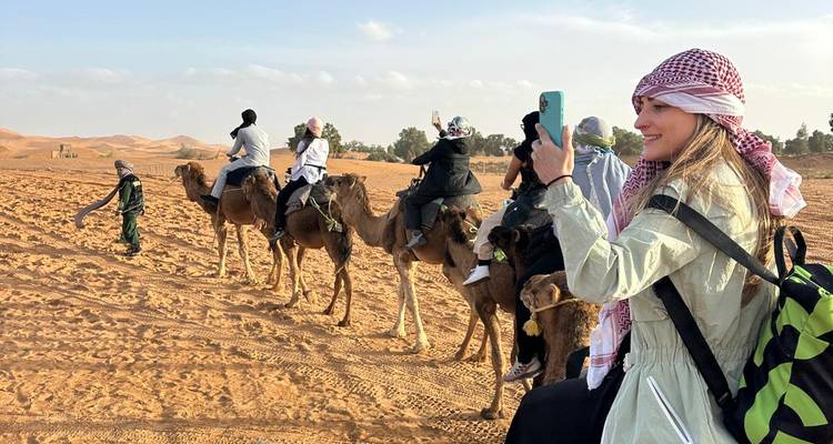 File de touristes sur des chameaux menée à travers le désert sablonneux pendant qu'un voyageur filme avec son téléphone.