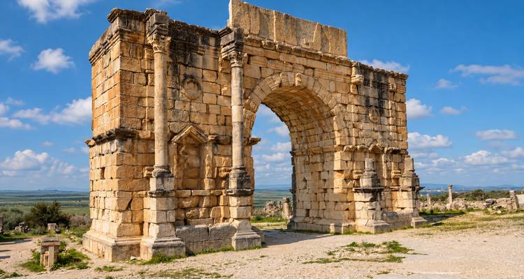 Un arco triunfal romano bien conservado se encuentra en un sitio arqueológico abierto bajo un cielo azul.