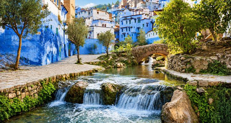 Un puente de piedra cruza un arroyo cristalino que cae en pequeñas cascadas junto a edificios de un azul vibrante.
