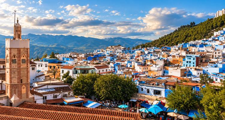 Paisaje panorámico de las casas azules de Chefchaouen cayendo en cascada por una ladera con telón de fondo montañoso y nubes dramáticas.