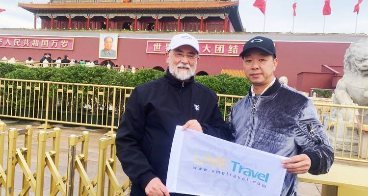 Two travellers pose with a tour banner in front of Tiananmen Gate decorated with red flags.