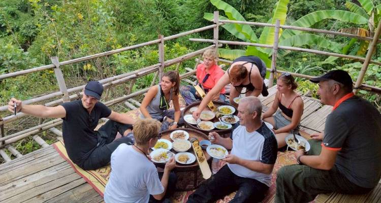 Los viajeros sentados en una cubierta de bambú disfrutan de un almuerzo tailandés compartido rodeados de follaje de la selva.