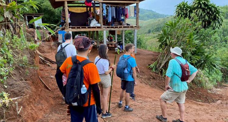 Grupo de senderistas hace una pausa en un sendero de tierra roja junto a una cabaña de madera sobre pilotes en las colinas.