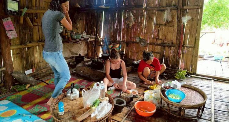 Los viajeros ayudan a preparar comida sobre un fuego de leña dentro de una cocina de aldea con paredes de bambú.