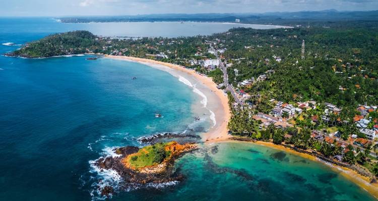 Aerial view of a curved tropical coastline with turquoise waters and a sandy beach town.