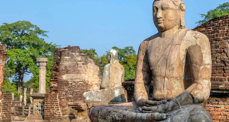 Ancient seated stone Buddha statue amid brick ruins and greenery under clear blue sky.