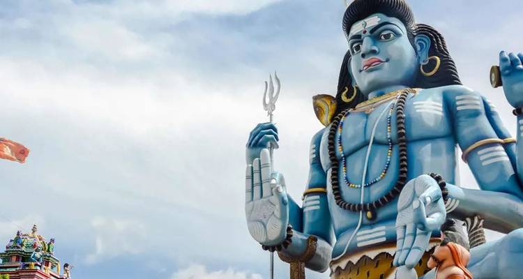 Riesige blaue Statue von Lord Shiva mit einem Dreizack erhebt sich gegen einen leicht bewölkten Himmel.