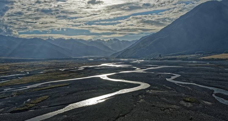 Des chenaux fluviaux tressés et réfléchissants serpentent à travers une large vallée alpine sous les imposants sommets de Nouvelle-Zélande.