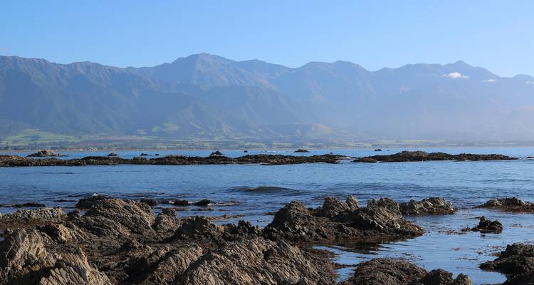 Des bassins rocheux de marée bordent une mer d'un bleu limpide avec la chaîne de montagnes de Kaikoura qui s'élève au loin.