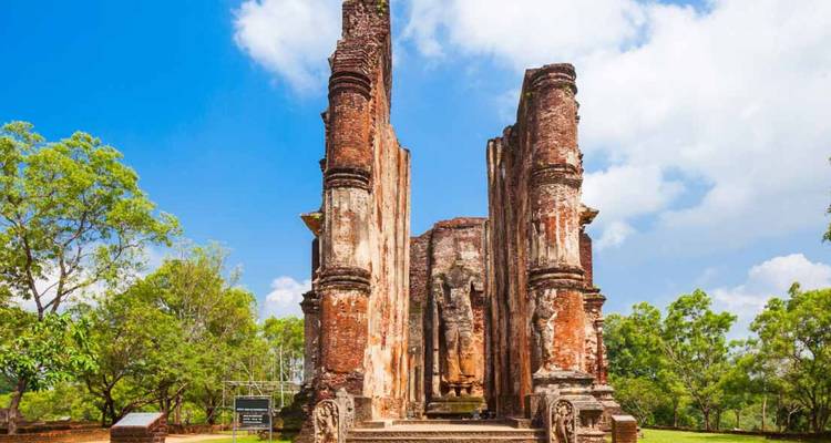 Ruined brick pillars and stone carvings of an ancient temple framed by lush greenery under a bright sky.