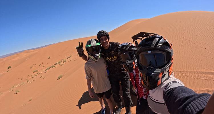 Quatre amis portant des casques de moto prennent un selfie sur une haute dune de sable orange sous un ciel bleu éclatant.