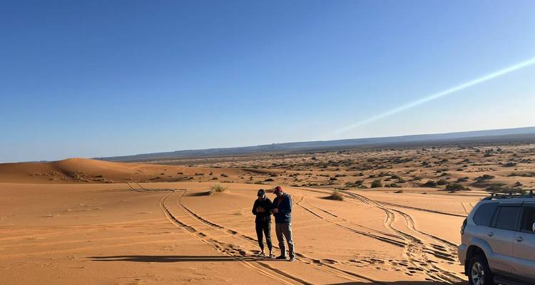 Deux voyageurs se tiennent à côté de traces de SUV dans un désert sablonneux ouvert avec un véhicule 4x4 garé à proximité.