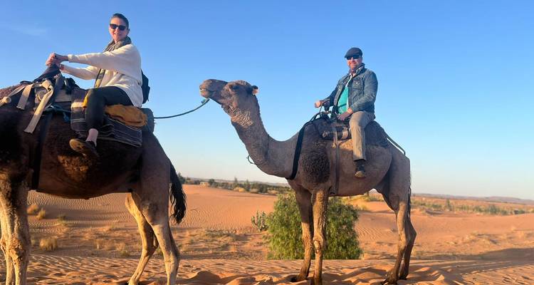 Deux touristes montent des chameaux à travers des dunes de sable orange ondulantes sous un ciel d'après-midi dégagé.
