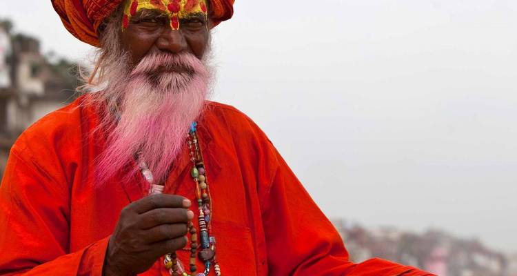 Sadhu pintado coloridamente con barba rosa vistiendo túnica naranja y collares de cuentas gesticulando con la mano