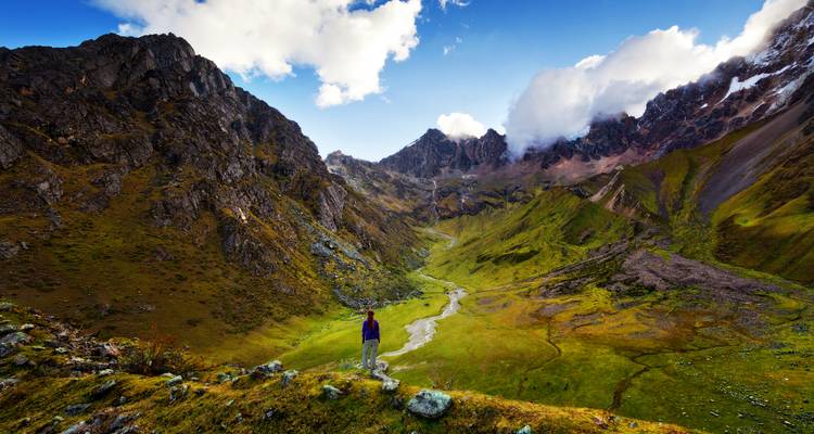 Randonneur solitaire contemplant une vallée alpine luxuriante entourée de pics andins escarpés et de nuages dramatiques
