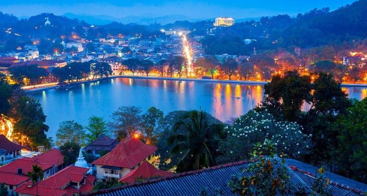 Abendstimmung über dem Kandy-See mit Stadtlichtern entlang der Uferpromenade und blauen Hügeln dahinter.