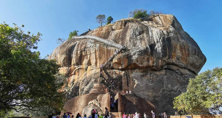Menschenmengen erklimmen die steile Treppe hinauf zum gigantischen Monolithen von Sigiriya unter klarem blauem Himmel.