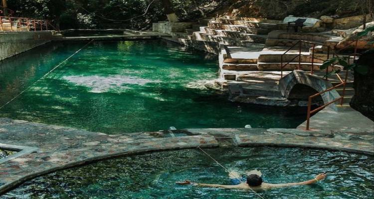 Person relaxing in a natural emerald-green hot spring pool surrounded by stone terraces