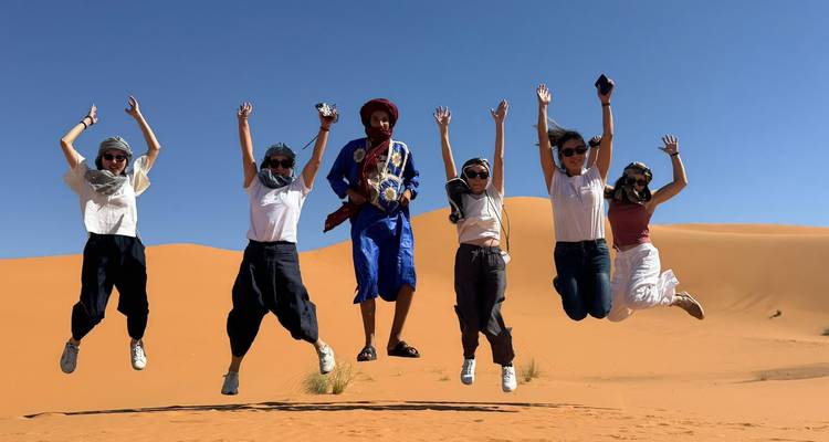 Groupe d'amis sautant joyeusement sur des dunes de sable orange sous un ciel bleu dégagé