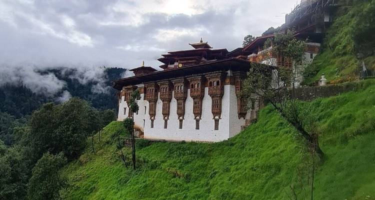 Remote Bhutanese monastery perched on a lush green mountainside with mist drifting through the valley.