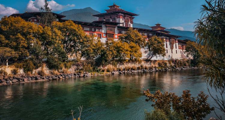 Punakha Dzong, gespiegelt in einem ruhigen Fluss und umrahmt von Herbstbäumen sowie entfernten, nebelverhangenen Bergen.