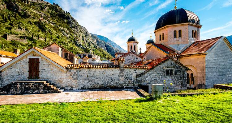 Iglesia de piedra con cúpulas oscuras ubicada entre montañas exuberantes y un cielo azul brillante en el casco antiguo de Kotor.