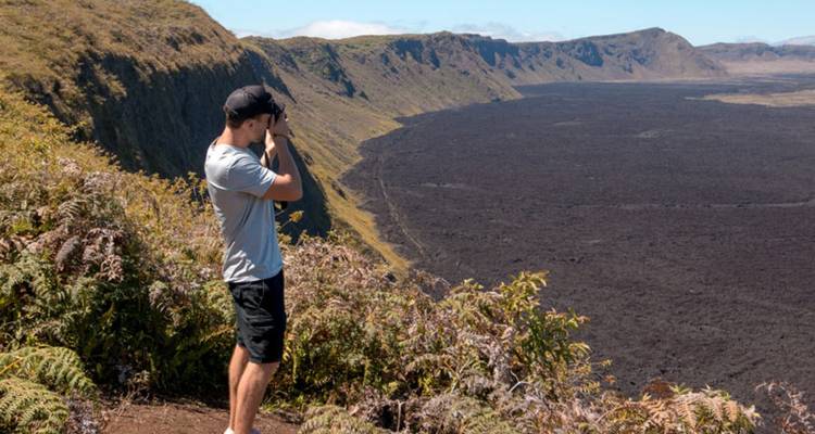Randonneur photographiant un vaste caldeira volcanique depuis un point de vue bordé de fougères luxuriantes.
