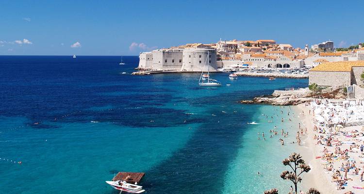 Kristalhelder turkoois water en strand met Dubrovnik's iconische ommuurde stad en vestingwerken die uitrijzen boven de Adriatische Zee.