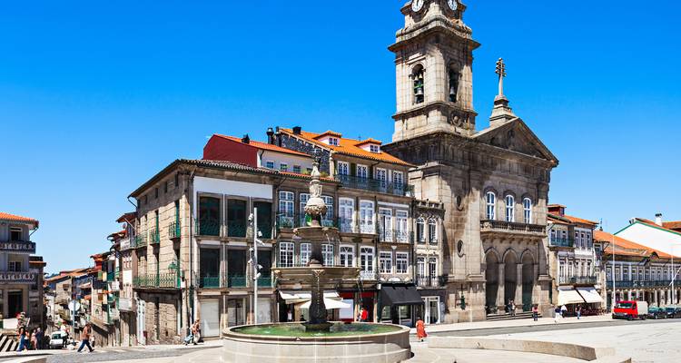 Una fuente de piedra histórica y una torre del reloj dominan una plaza soleada en el casco antiguo de Guimarães.