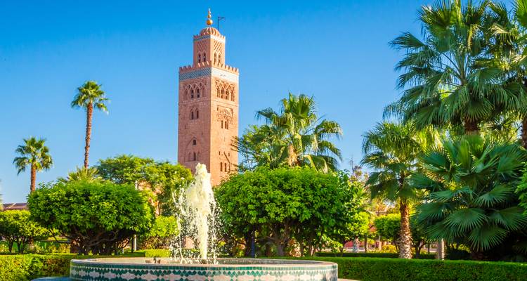 Vue de la mosquée de la Koutoubia s'élevant derrière une fontaine carrelée et un jardin luxuriant bordé de palmiers sous un ciel bleu dégagé.