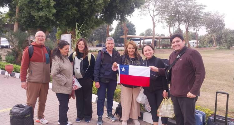Grupo de viajeros posando al aire libre con equipaje y una bandera chilena en un punto de encuentro rodeado de vegetación.