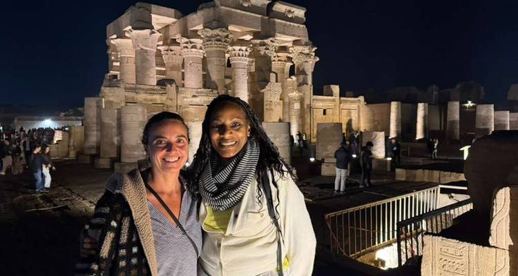 Dos mujeres sonríen frente a un templo egipcio antiguo dramáticamente iluminado por la noche.