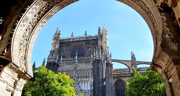 Ein kunstvoll geschnitzter maurischer Bogen rahmt den aufragenden gotischen Turm der Kathedrale von Sevilla vor blauem Himmel ein.