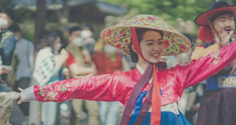 Mujer sonriente en hanbok colorido y sombrero tradicional realizando una danza con multitud borrosa detrás.
