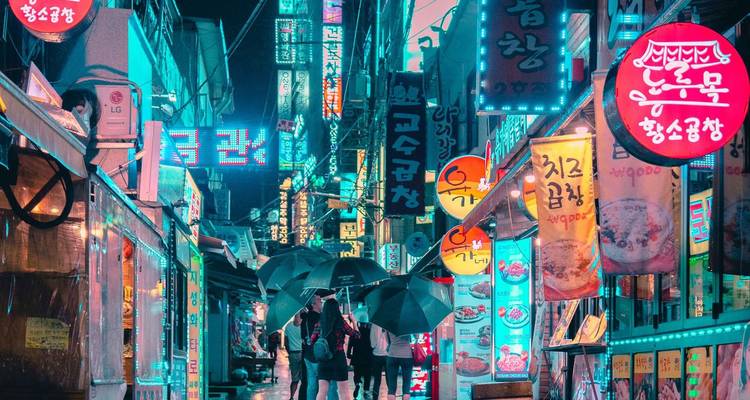 Callejón vibrante iluminado con neón en Seúl de noche con gente caminando bajo paraguas negros entre carteles coloridos.