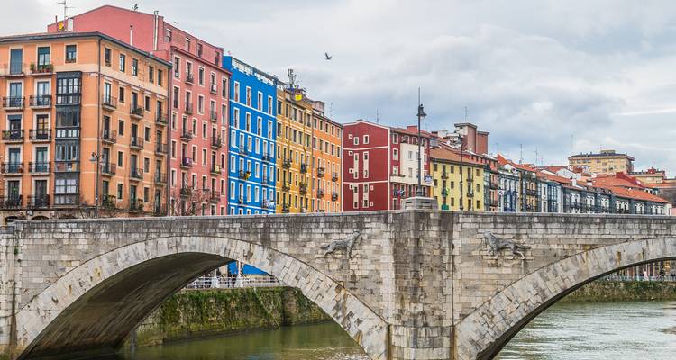 Des façades d'appartements colorées surplombent la rivière Nervión derrière un vieux pont de pierre par une journée nuageuse à Bilbao.
