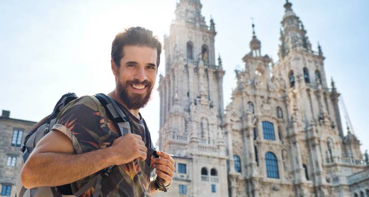 Un randonneur souriant pose devant les tours jumelles ornées de la cathédrale de Saint-Jacques-de-Compostelle éclairées par le soleil matinal.