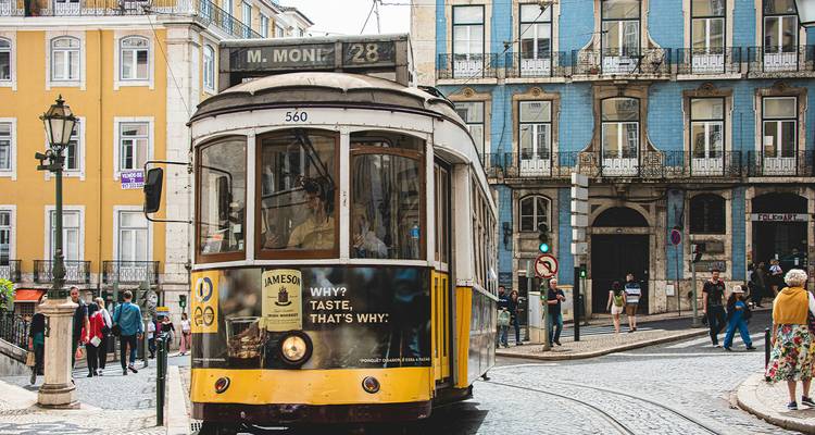 Tramway jaune classique 28 naviguant dans une rue pavée de Lisbonne bordée de bâtiments colorés et de piétons.