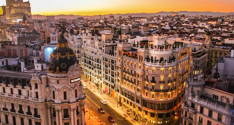 Vue aérienne au coucher du soleil le long de la Gran Vía de Madrid avec des toits ornés et des éclairages de rue lumineux s'étendant vers les montagnes lointaines.