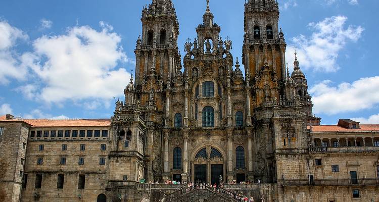 Grande façade baroque de la cathédrale de Saint-Jacques-de-Compostelle avec des foules rassemblées sur l'escalier de pierre sous un ciel lumineux.