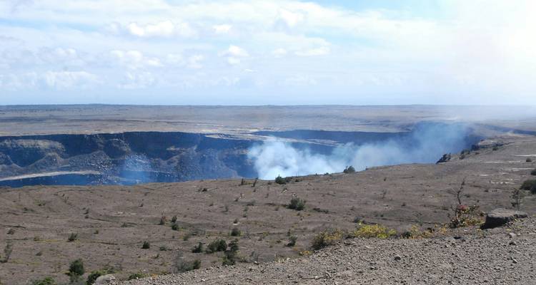 Cratère volcanique fumant situé dans un vaste champ de lave stérile sous un ciel brumeux.