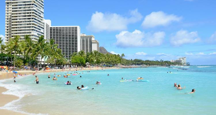 Plage animée de Waikiki avec nageurs et baigneurs de soleil, bordée par des hôtels de grande hauteur et une eau bleu clair.