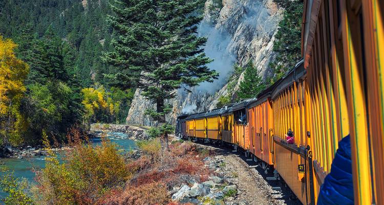 Histórico tren amarillo bordeando un acantilado sobre un río turquesa entre follaje otoñal en Colorado.