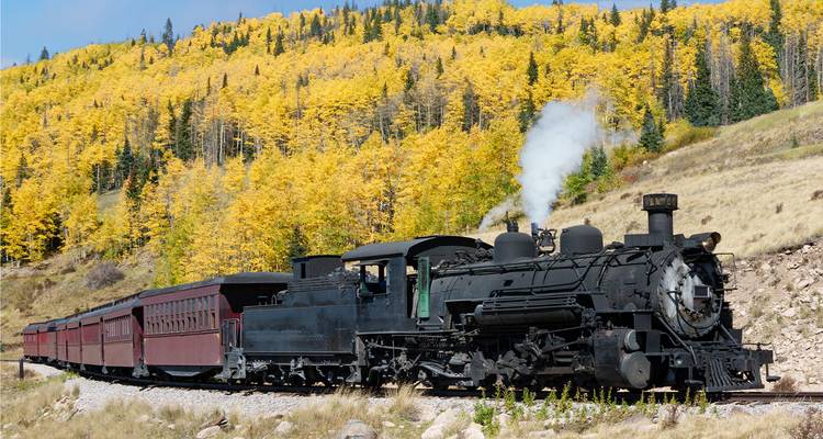 Locomotora de vapor negra tirando vagones rojos pasando por bosques dorados de álamos temblones en una vía de montaña.