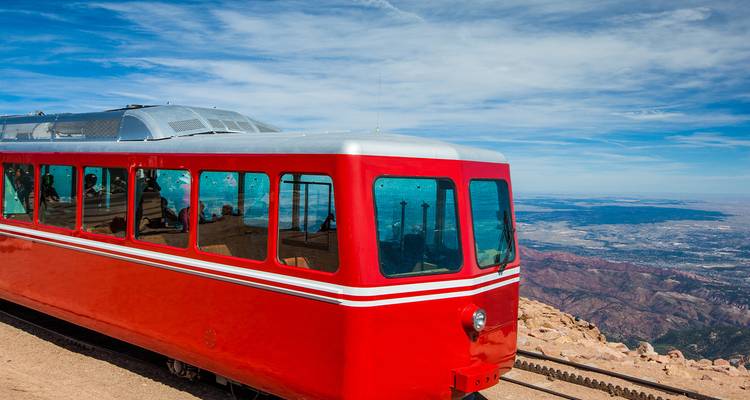 Vagón rojo brillante del ferrocarril cremallera de Pikes Peak posado en la cima de una montaña con vastas llanuras abajo.