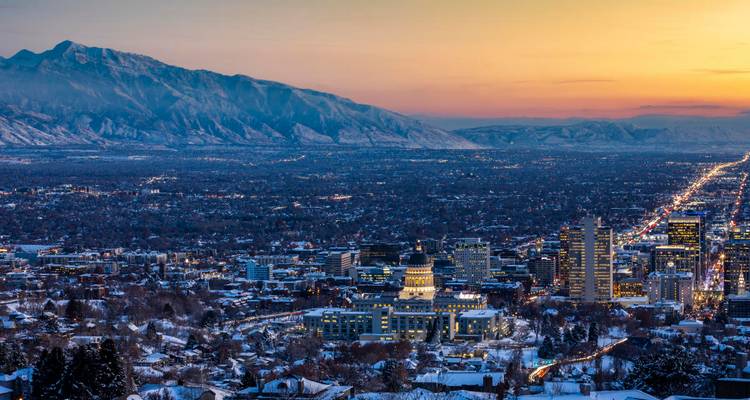 Vista amplia del atardecer sobre Salt Lake City con el edificio del Capitolio iluminado y el telón de fondo de montañas cubiertas de nieve.